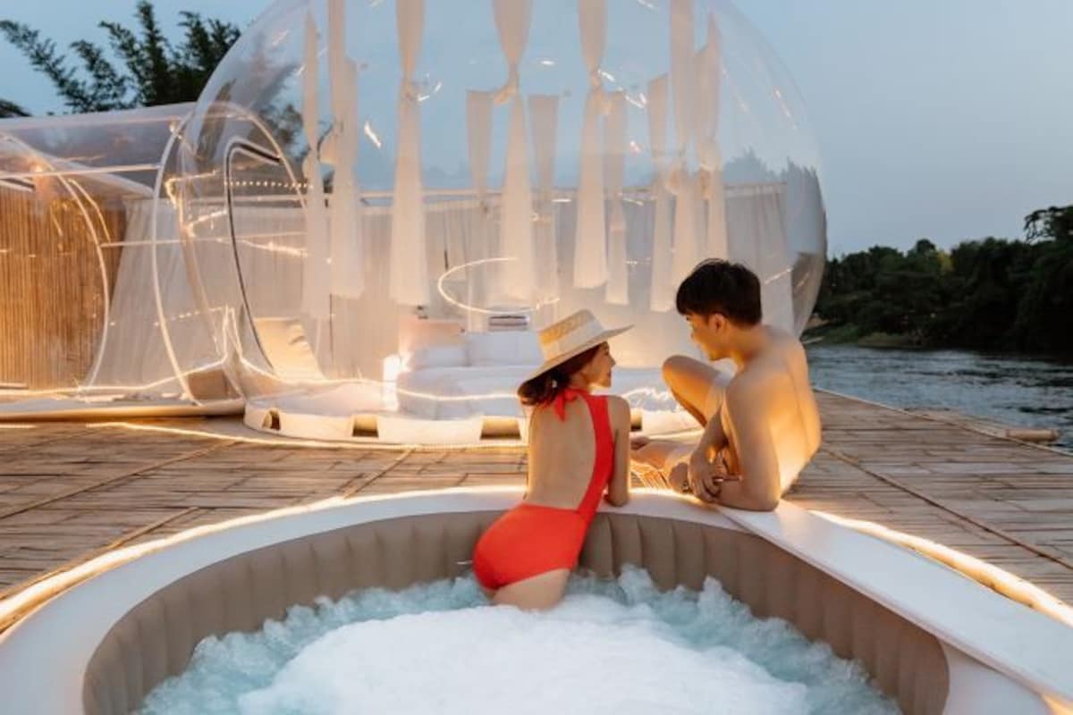 Una pareja relajándose en una bañera de burbujas transparente rodeada por una terraza de madera y un río.