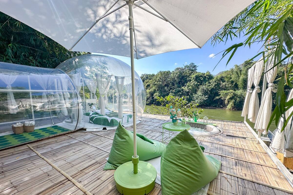 A transparent greenhouse structure with a bed and seating area, surrounded by bamboo flooring and green cushions.