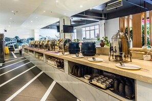 Ein reichhaltiges Buffet mit verschiedenen Speisen und Getränken, eine Sitzgelegenheit mit Stühlen und eine Theke mit einer Kaffeemaschine.