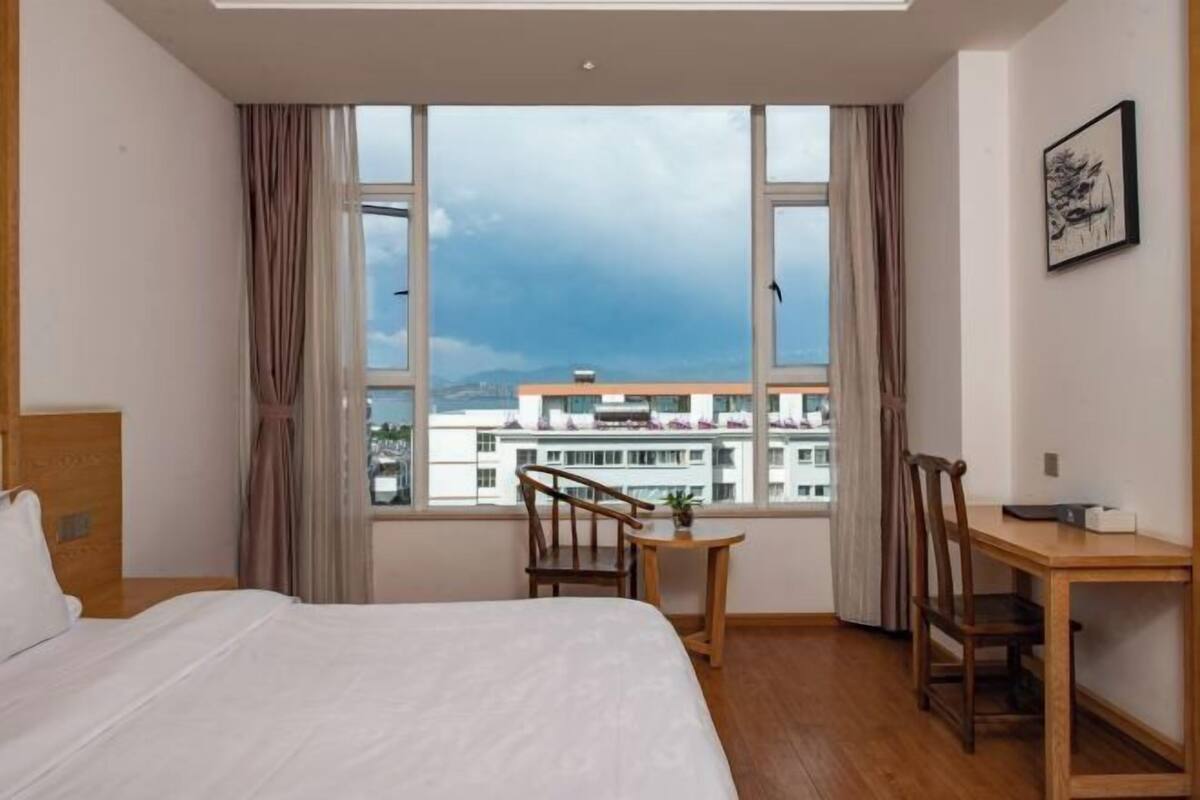 A hotel room with a bed, a desk, a chair, a small table, and a window with a view of buildings and mountains.