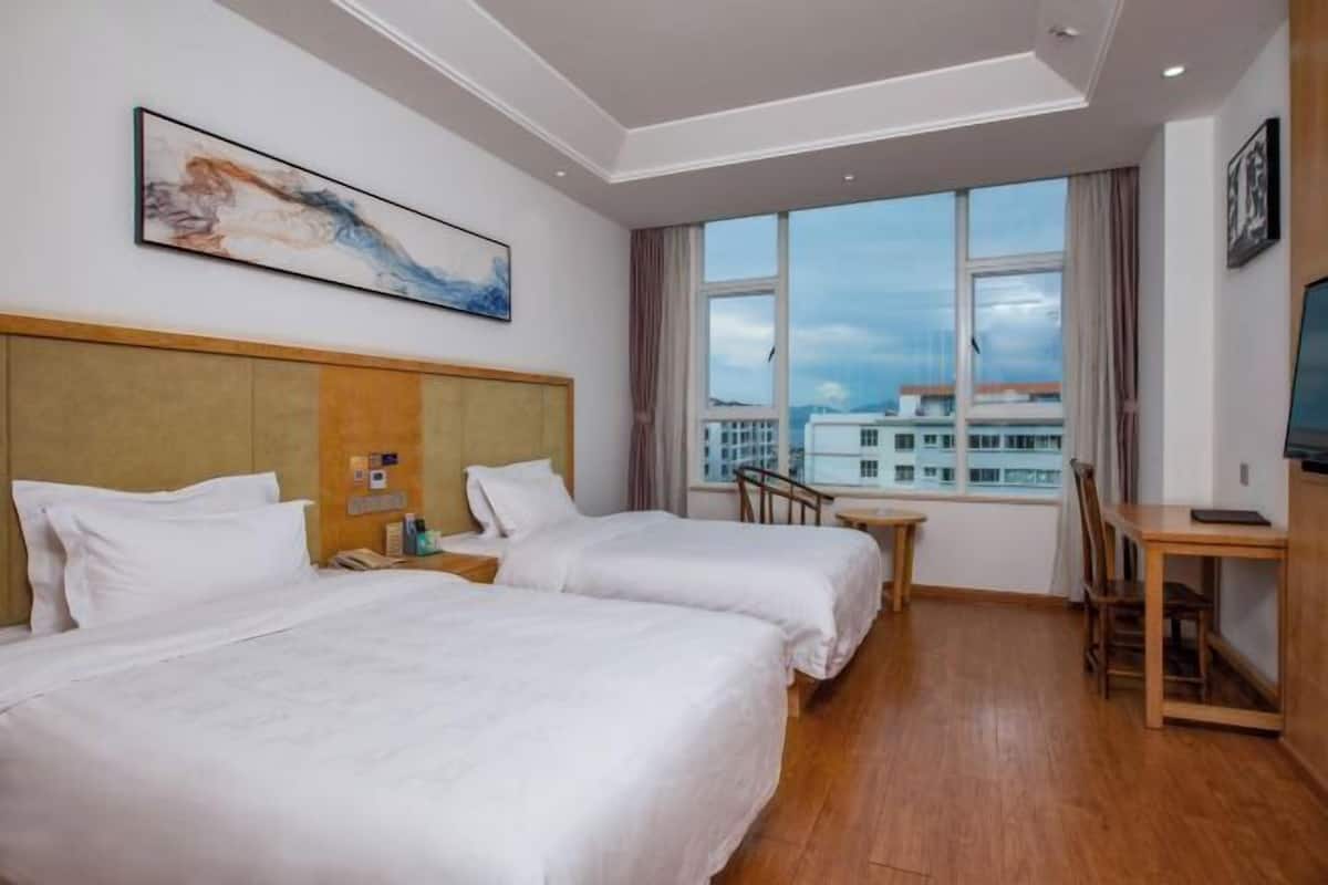 A hotel room with two beds, a desk, a chair, a TV, and a large window with a city view.