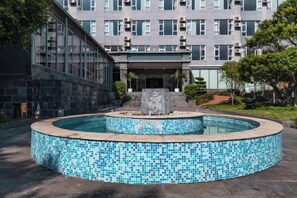 Una piscina con borde de azulejos, una escultura de piedra y un edificio con múltiples ventanas al fondo.