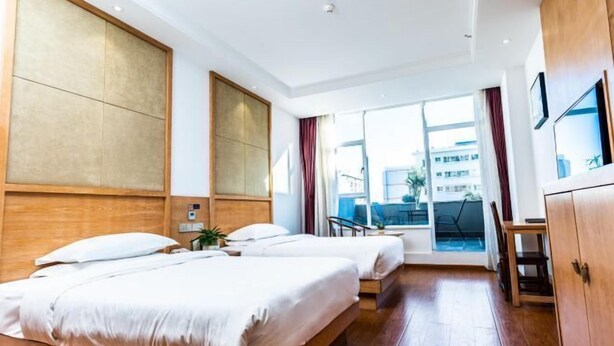 A hotel room with two beds, a wooden headboard, a large window with curtains, a desk, and a view of buildings outside. - Dali Wenyue Hotel (Erhai Ecological Corridor Branch) (Dali City)