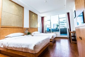 Ein Hotelzimmer mit zwei Betten, einem Holzkopfende, einem großen Fenster mit Vorhängen, einem Schreibtisch und Blick auf Gebäude.