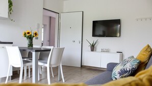 Un salon moderne avec un canapé gris, une table à manger blanche avec des chaises, un téléviseur à écran plat fixé au mur et une petite plante en pot posée sur un meuble blanc.