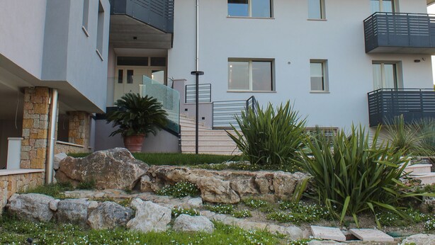 Un bâtiment moderne doté de nombreux balcons, d’une façade en pierre et d’un jardin aménagé avec des rochers et des plantes.
