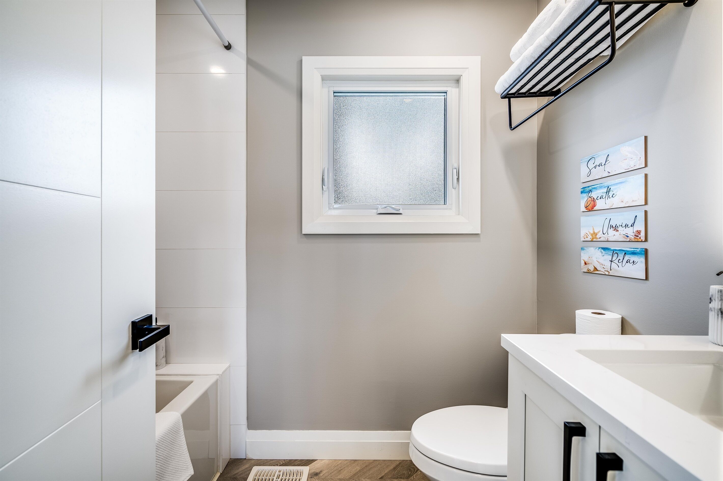 A modern bathroom with a bathtub, toilet, and sink. A window with frosted glass is above the toilet, and there are three framed pictures on the wall.