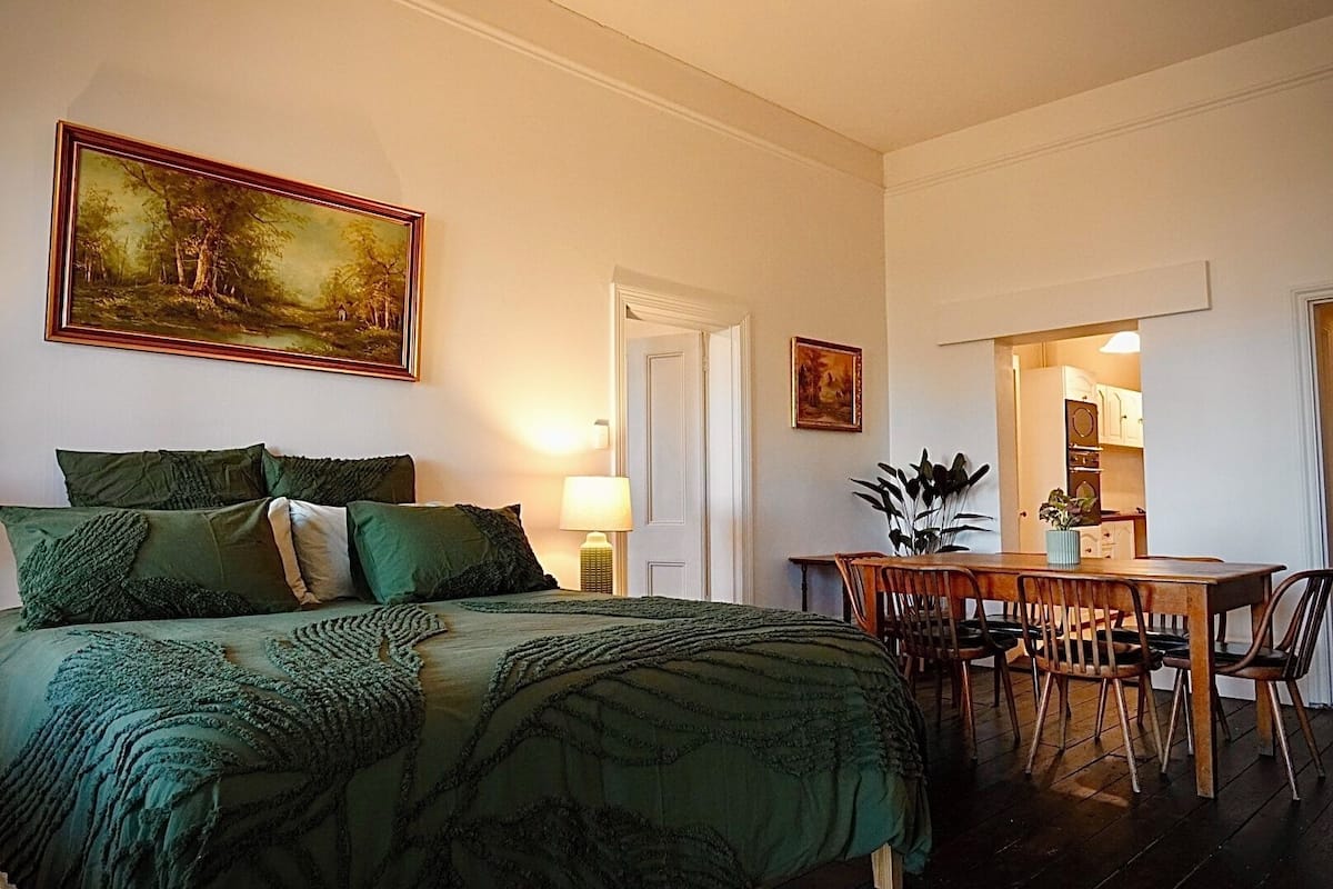 A bedroom with a bed, green bedding, a painting, a desk, and a chair.