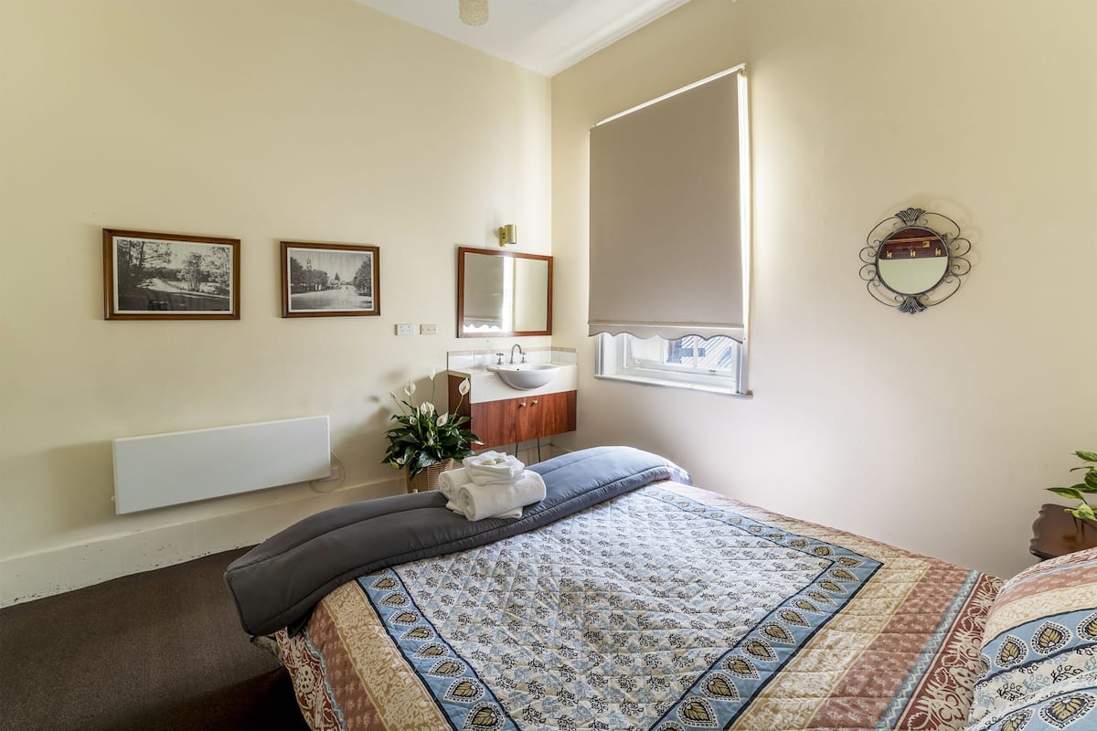 A bedroom with a bed, a bedside table with a lamp, a window with a view, and a bathroom with a sink and mirror.