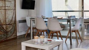 Un espace repas moderne avec une table en verre, des chaises blanches et un téléviseur fixé au mur.