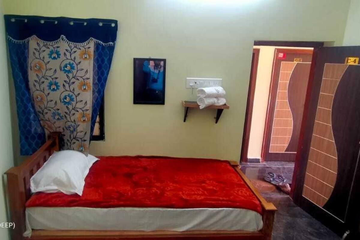 A single bed with a red blanket, a wall-mounted shelf with towels, a door with a number, and a curtain with a floral pattern.