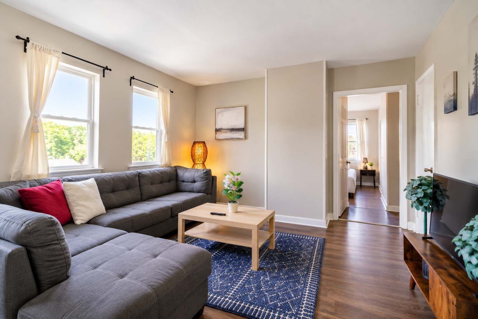 A modern living room with a grey sofa, a wooden coffee table, and a blue rug. There is a window with curtains, a lamp, and a plant on the side.