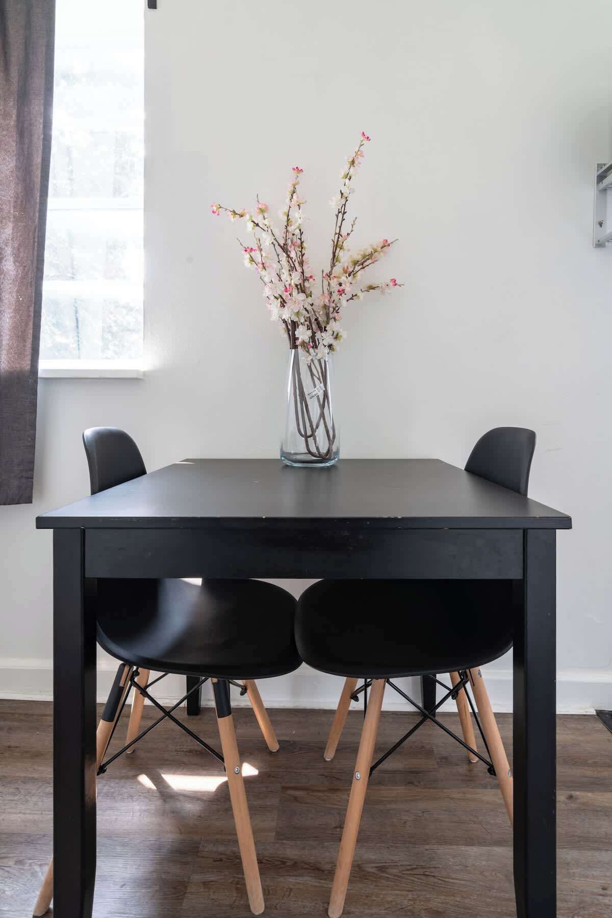 Une table à manger avec deux chaises noires, un vase avec des fleurs et une fenêtre avec des rideaux.