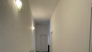 Un couloir étroit aux murs blancs et au sol carrelé. Un radiateur se trouve sur la gauche et une porte se trouve au fond du couloir.