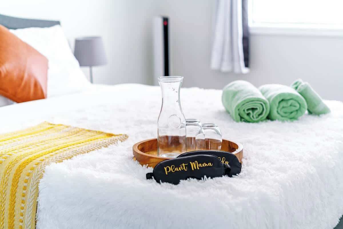 A bed with a white fluffy blanket, a yellow and white striped pillow, a black pillow with "Plant Mama" text, a wooden tray with a glass bottle, and green towels.