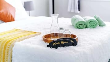 A bed with a white fluffy blanket, a yellow and white striped pillow, a black pillow with "Plant Mama" text, a wooden tray with a glass bottle, and green towels.