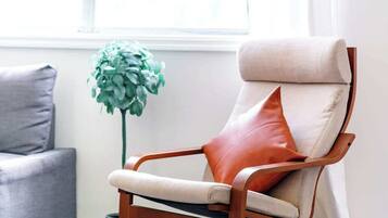 A modern living room with a light-colored sofa, a wooden rocking chair with a beige cushion and a red pillow, and a green plant.