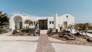 A white building with a central entrance, surrounded by a gravel path and various plants. - Echo Villa (Santorini)