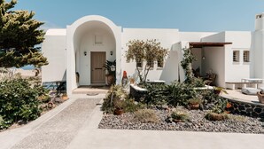 A white building with an arched entrance, a stone pathway, and a garden with various plants and a small waterfall. - Echo Villa (Santorini)