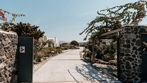 A paved pathway leading to a stone gate with a sign, surrounded by greenery and buildings in the background. - Echo Villa (Santorini)