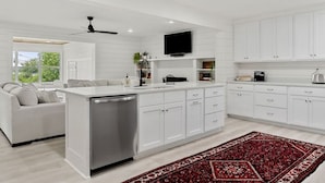 A modern kitchen with white cabinetry, a built-in dishwasher, a flat-screen TV, and a red patterned rug. - Sunset Cove- Lake Home, Dock, Fire Pit, Hot Tub, Game Room, Etc (Winchester)