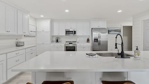 A modern kitchen with white cabinetry, stainless steel appliances, a large island, and a bar area with chairs. - Sunset Cove- Lake Home, Dock, Fire Pit, Hot Tub, Game Room, Etc (Winchester)