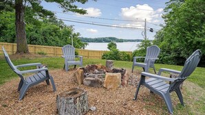 A patio with four Adirondack chairs arranged around a wood fire pit, overlooking a lake. - Sunset Cove- Lake Home, Dock, Fire Pit, Hot Tub, Game Room, Etc (Winchester)
