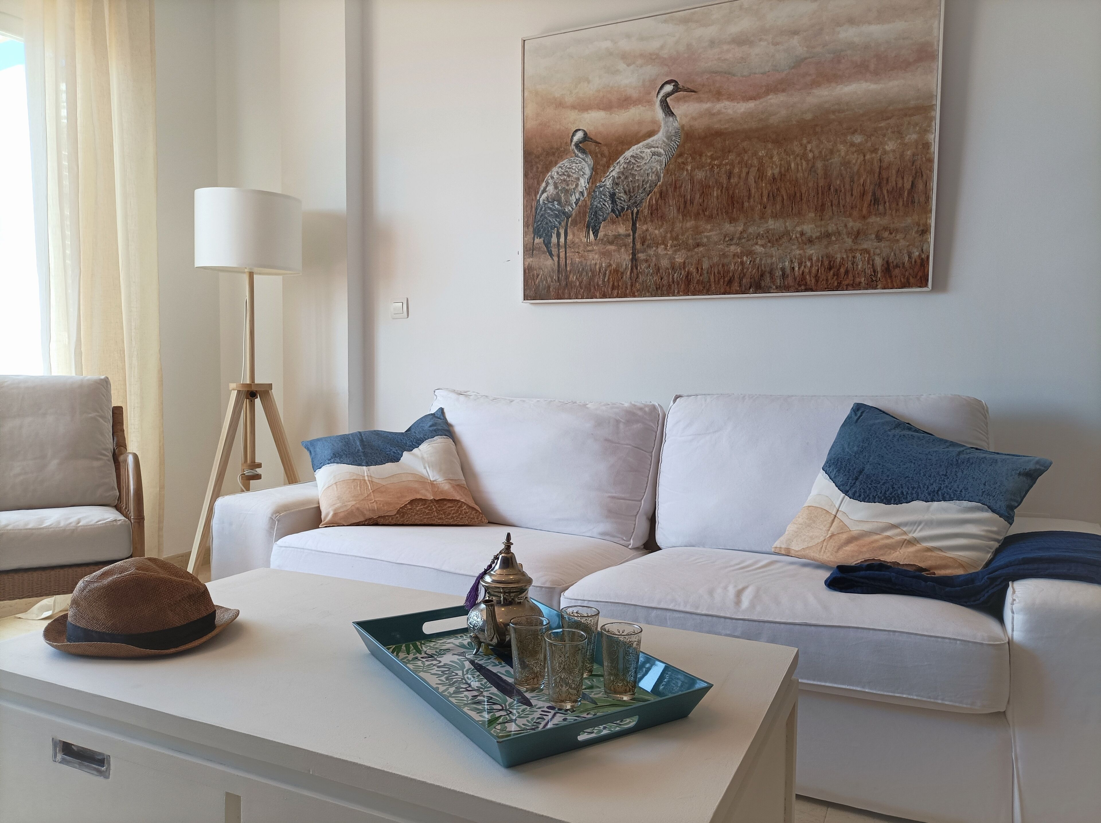 A living room with a white sofa, a coffee table with a tray, a lamp, and a painting of birds on the wall.