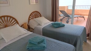 A hotel room with two beds, each with a blue bedspread and a folded towel. There is a wicker chair and a small table with a lamp.