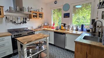 Een keuken met een fornuis, spoelbak en een houten aanrecht. Er zijn kastjes, een klok en een raam met uitzicht op bomen.