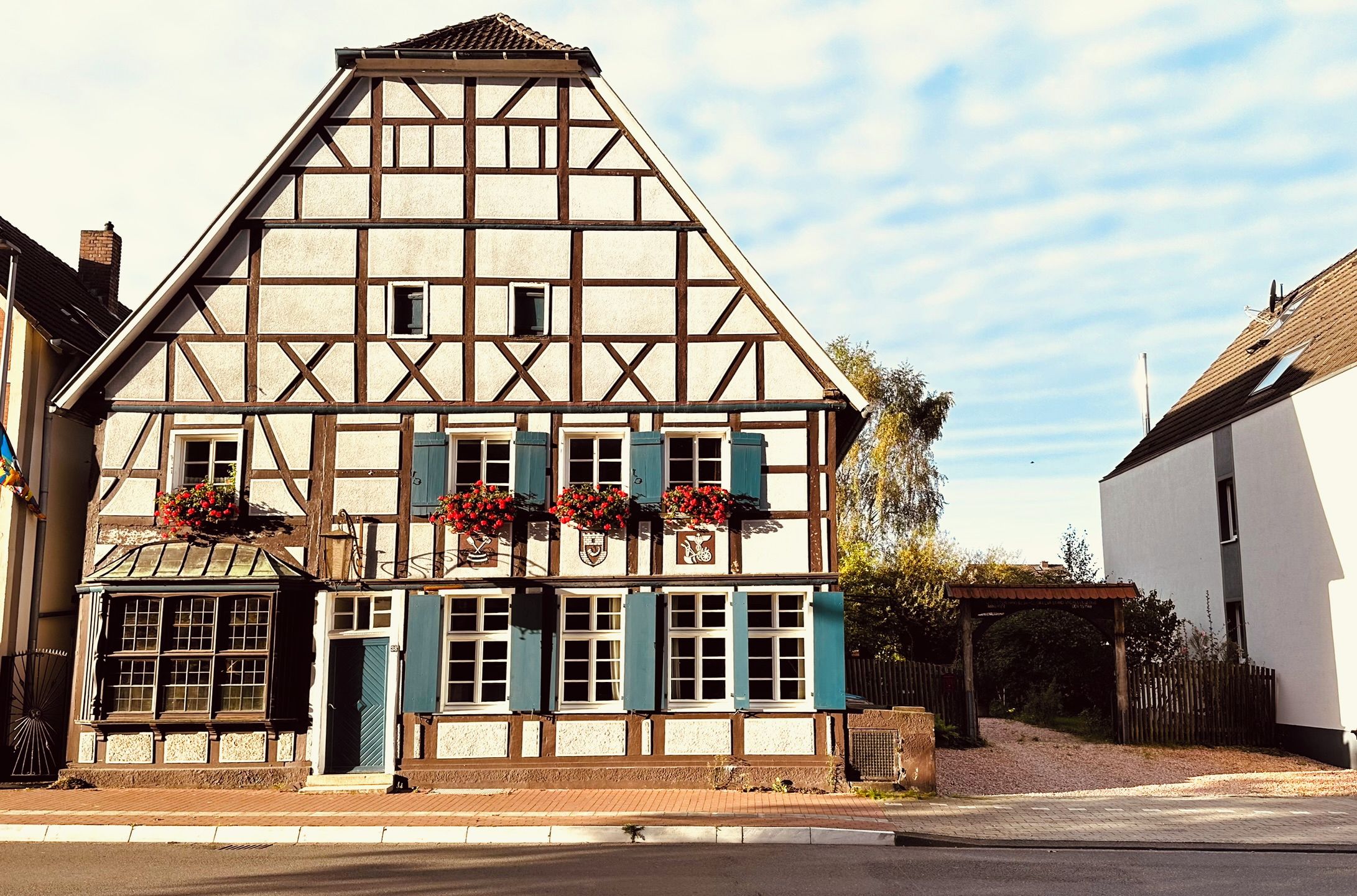 Un bâtiment traditionnel à colombages, doté de nombreuses fenêtres et volets, d’un petit porche et de jardinières fleuries.