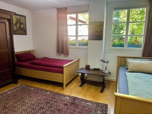 Een kamer met een bed, een bureau met een lamp, een raam met uitzicht op groen, en een kamerkleed met een patroon.