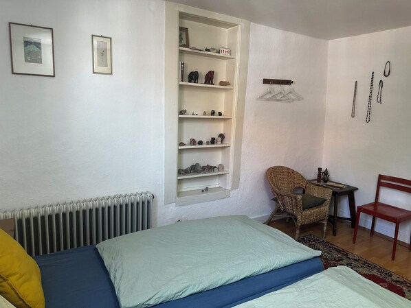 A bedroom with a bed, a chair, a shelf with figurines, and a radiator.