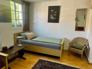 A compact bedroom with a single bed, a desk with a lamp, a chair, a mirror, and a rug.