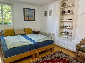 A bedroom with a double bed, a radiator, a shelf with figurines, and a window with a view of greenery. - Historic half-timbered house in Westphalia (Datteln)
