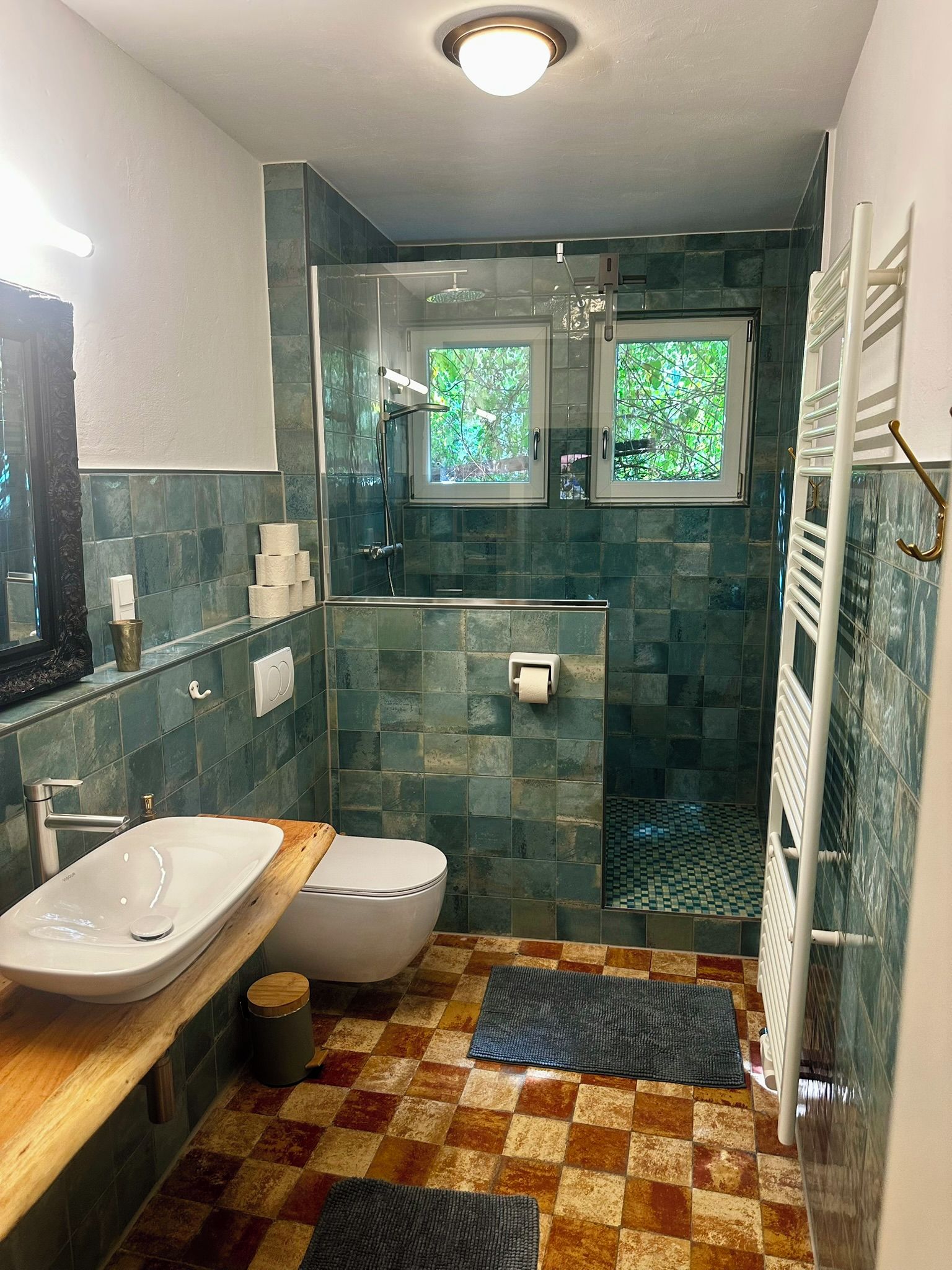 Une salle de bain équipée d’une douche, de toilettes et d’un lavabo, avec un carrelage à motifs et une fenêtre donnant sur les arbres.