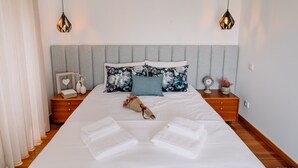 A neatly made bed with white linens, a floral pillow, and a small bouquet of flowers. There are two bedside tables with decorative items and a wall-mounted air conditioner above the bed.
