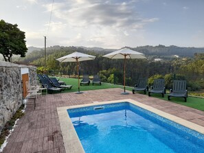 A swimming pool with lounge chairs and umbrellas, surrounded by a brick deck and a view of a hilly landscape. - Coliving The VALLEY Portugal countryside retreat for digital nomads and remote workers (Vale de Cambra)