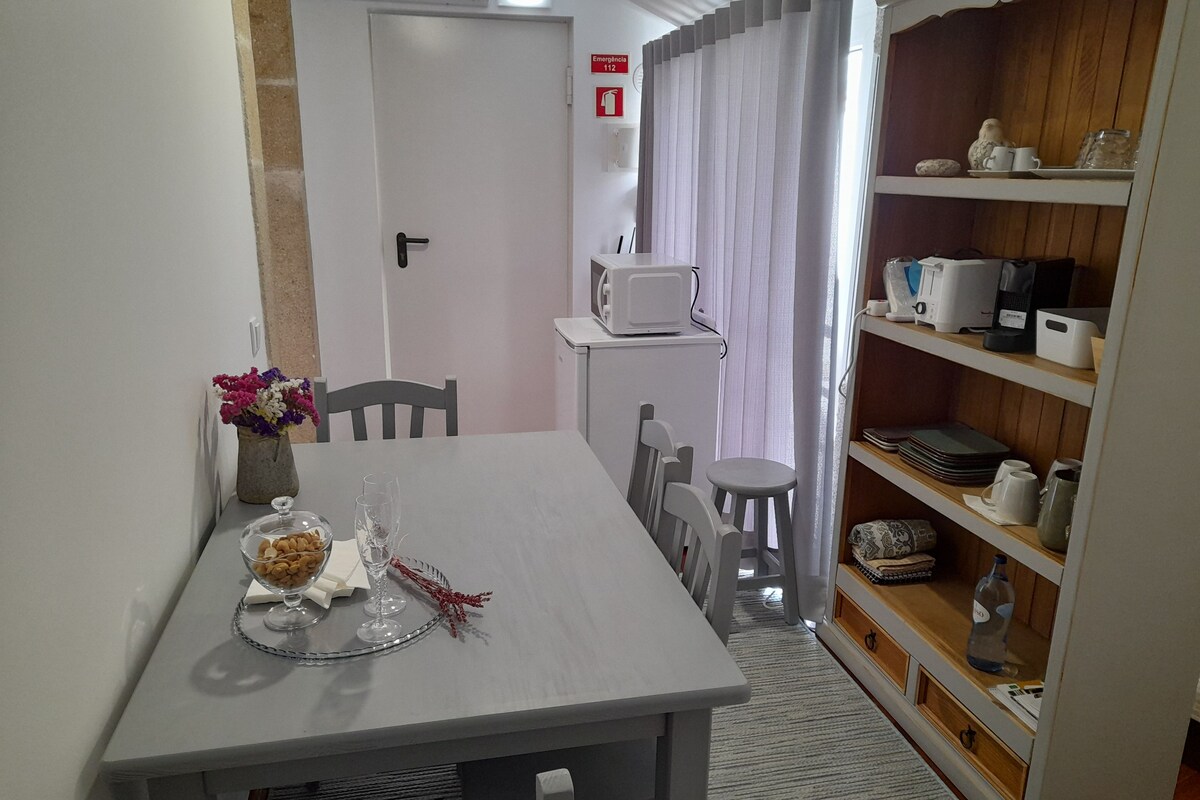 A dining area with a table set for two, a microwave, and a shelf with kitchen items.