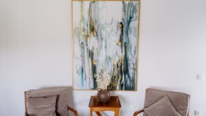 A room with two wooden chairs, a small wooden table with a vase, and a large abstract painting on the wall.