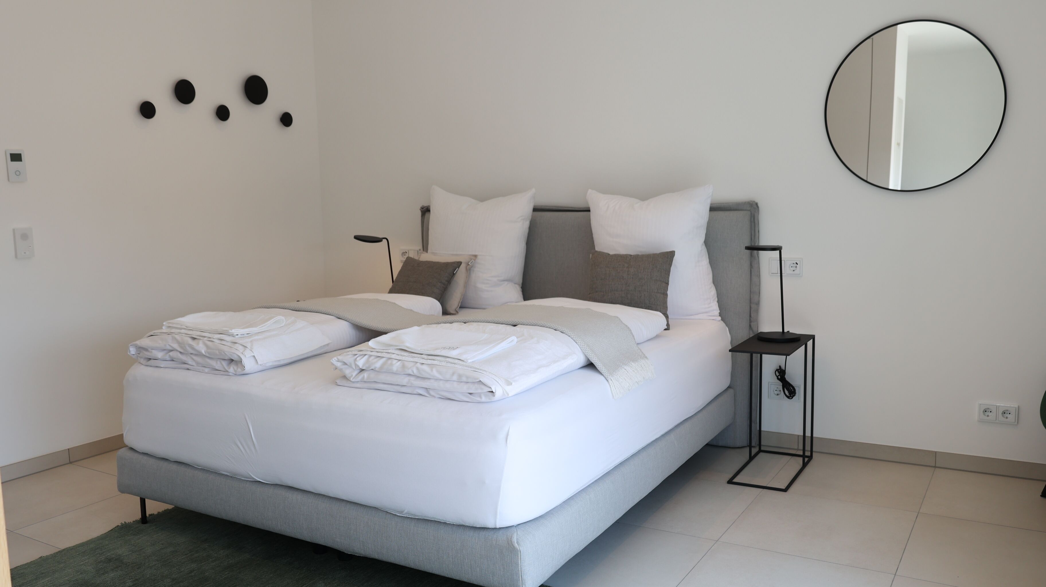 A modern bedroom with a bed, pillows, a bedside table, a lamp, and a wall-mounted mirror.