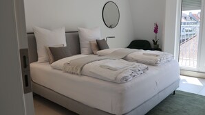 Una cama bien tendida con sábanas blancas y cabecera gris, un espejo redondo en la pared, y vistas a un balcón con barandilla.