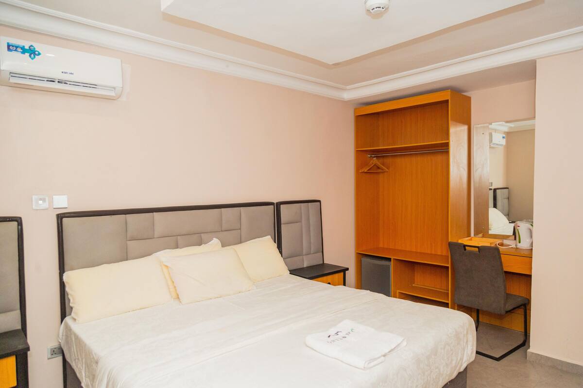 A hotel room with a bed, a desk, a chair, a wardrobe, and an air conditioning unit.