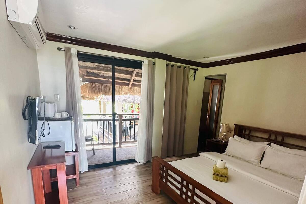 A hotel room with a wooden bed, a small desk with a chair, a TV, and a balcony with a thatched roof.
