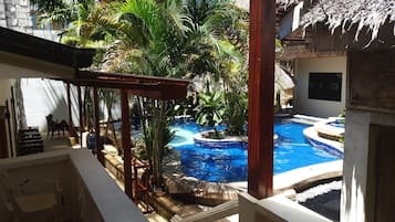 A pool area with a thatched roof structure, surrounded by lush greenery and palm trees.