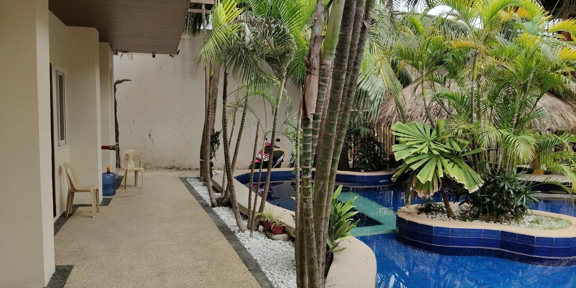A narrow corridor with a pool area featuring palm trees and a thatched-roof structure.