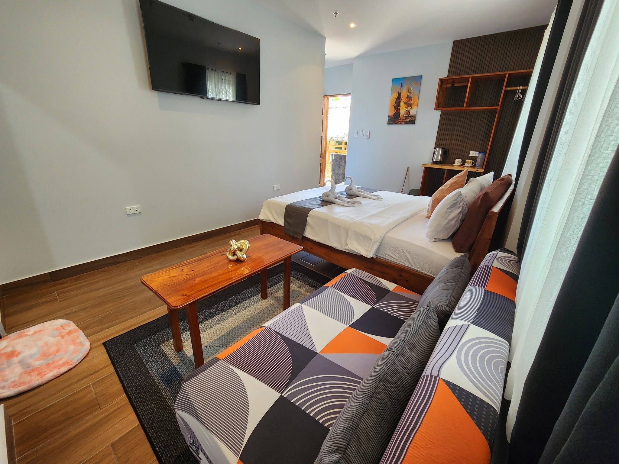 A compact hotel room with a bed, a sofa, a small table, and a TV mounted on the wall.