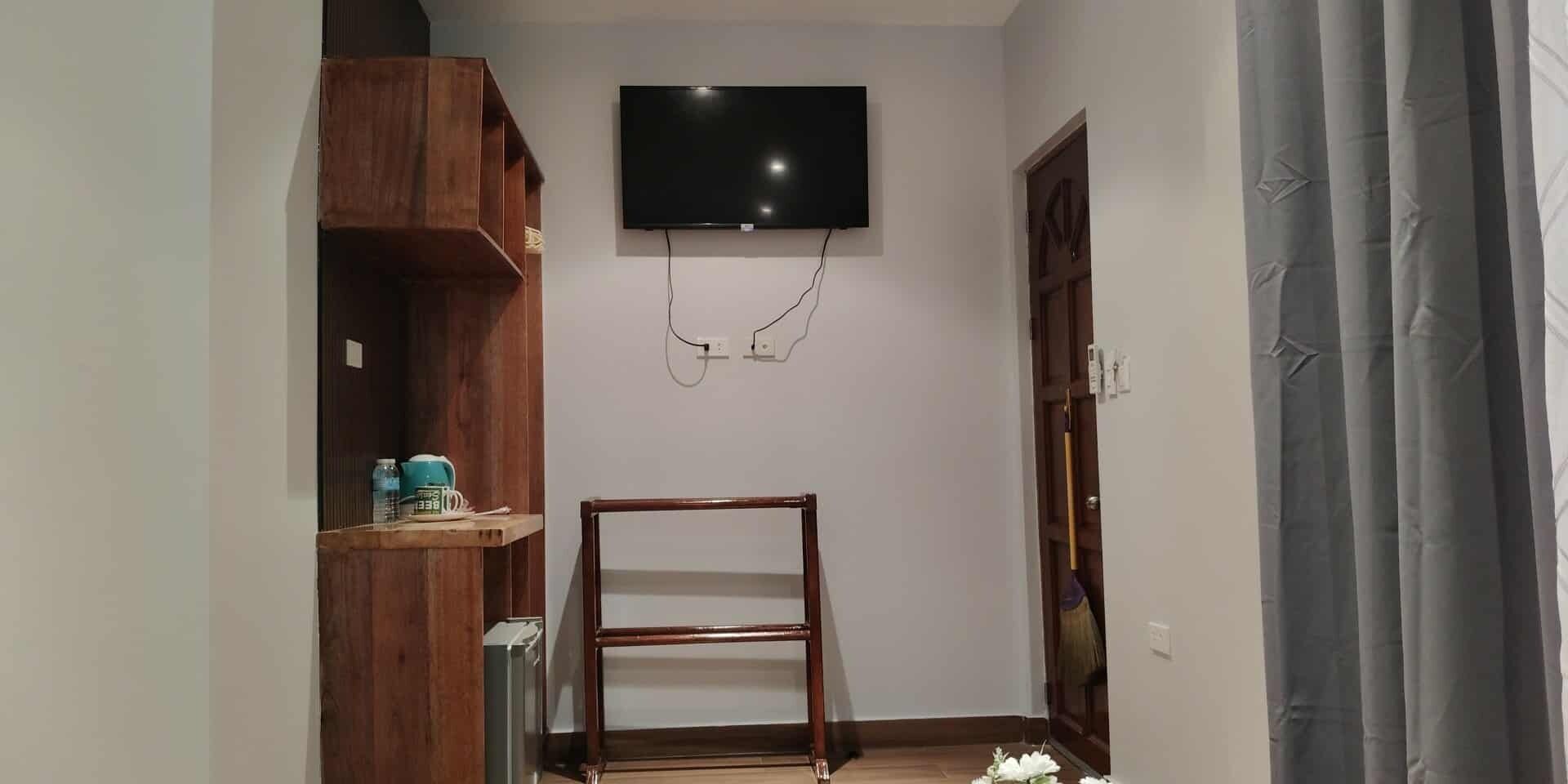 A room with a TV mounted on the wall, a wooden shelf with items, and a door with a broom leaning against it.