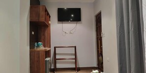 A room with a TV mounted on the wall, a wooden shelf with items, and a door with a broom leaning against it. - Alona Beach Poolside Hotel (Panglao)