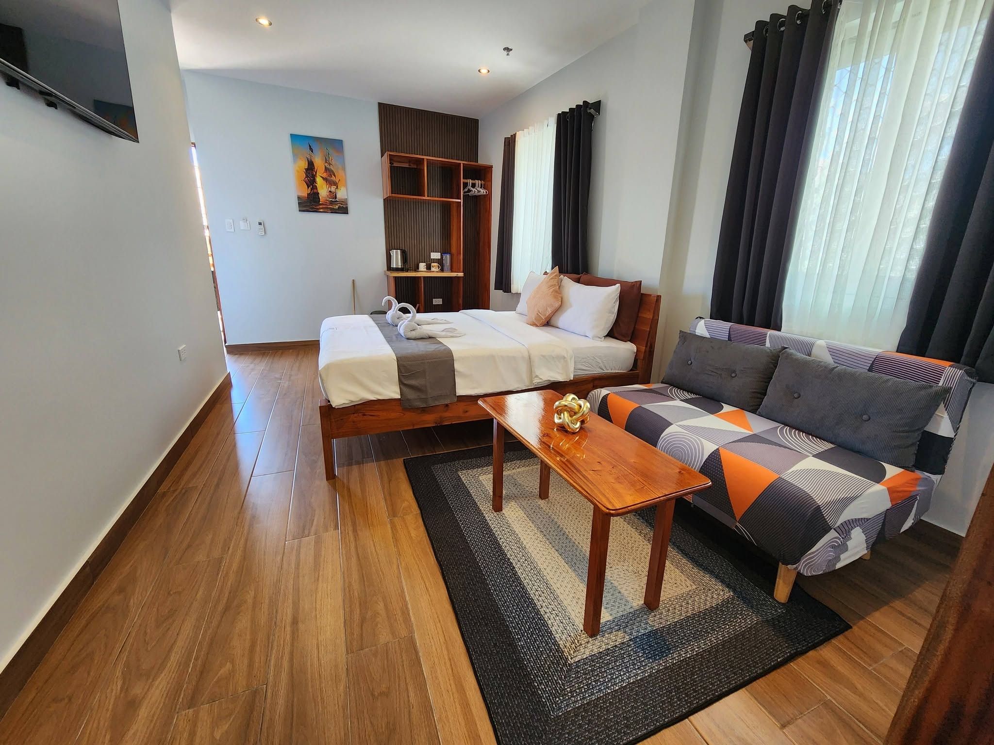 A hotel room with a bed, a sofa, a wooden table, a painting on the wall, and a window with curtains.
